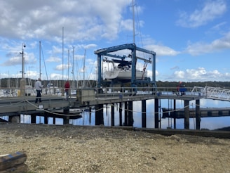 A boat is being lifted by a large crane-like structure on a marina dock. Several people are walking and standing on the dock, observing the process. The background features numerous sailboat masts and a body of water reflecting the cloudy blue sky.