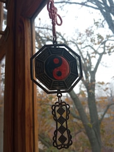 A hanging decoration with a Yin-Yang symbol inside an octagonal frame is suspended in front of a window. The background features a view of bare trees, suggesting a winter or late autumn setting. The wooden frame of the window is visible, adding a rustic feel.