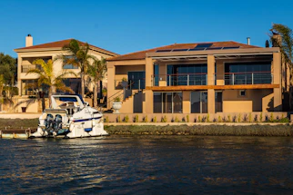 A modern waterfront house features large windows and a balcony with solar panels on the roof. Palm trees are planted in front, enhancing the tropical atmosphere. A docked motorboat is present in the adjacent waterway.