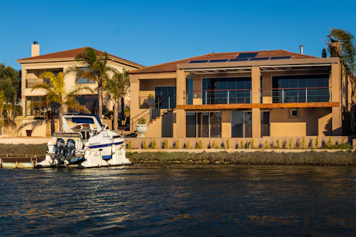 A modern waterfront house features large windows and a balcony with solar panels on the roof. Palm trees are planted in front, enhancing the tropical atmosphere. A docked motorboat is present in the adjacent waterway.