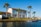 A two-story house with a red-tiled roof is situated by a waterfront. The house has large windows and a patio area. The surrounding area is landscaped with green grass and several tall palm trees. There is a small dock or pier extending into the water and the sky is clear and blue.