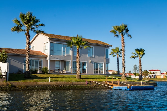 A two-story house with a red-tiled roof is situated by a waterfront. The house has large windows and a patio area. The surrounding area is landscaped with green grass and several tall palm trees. There is a small dock or pier extending into the water and the sky is clear and blue.