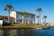 A two-story house with a red-tiled roof is situated by a waterfront. The house has large windows and a patio area. The surrounding area is landscaped with green grass and several tall palm trees. There is a small dock or pier extending into the water and the sky is clear and blue.