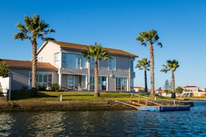 A two-story house with a red-tiled roof is situated by a waterfront. The house has large windows and a patio area. The surrounding area is landscaped with green grass and several tall palm trees. There is a small dock or pier extending into the water and the sky is clear and blue.