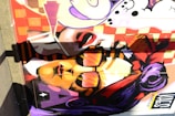 A vibrant mural features a stylized portrait of a person wearing sunglasses with a dynamic expression. The background consists of bold patterns and geometric shapes, with a mix of bright colors including orange, red, and purple. The artwork incorporates various graphic elements and text.