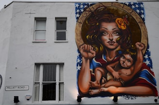 A mural on a building wall depicts a woman with long hair, holding a child. They both have raised fists, symbolizing empowerment and strength. The woman has a flower in her hair and a vibrant shawl with a pattern resembling the American flag. Above them, the word 'EQUALITY' is inscribed within a circular frame.