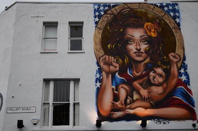 A mural on a building wall depicts a woman with long hair, holding a child. They both have raised fists, symbolizing empowerment and strength. The woman has a flower in her hair and a vibrant shawl with a pattern resembling the American flag. Above them, the word 'EQUALITY' is inscribed within a circular frame.