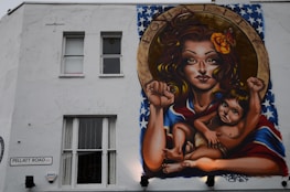 A mural on a building wall depicts a woman with long hair, holding a child. They both have raised fists, symbolizing empowerment and strength. The woman has a flower in her hair and a vibrant shawl with a pattern resembling the American flag. Above them, the word 'EQUALITY' is inscribed within a circular frame.