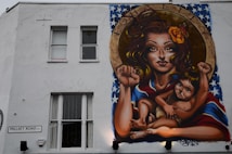 A mural on a building wall depicts a woman with long hair, holding a child. They both have raised fists, symbolizing empowerment and strength. The woman has a flower in her hair and a vibrant shawl with a pattern resembling the American flag. Above them, the word 'EQUALITY' is inscribed within a circular frame.