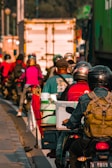 Several motorcyclists are riding together closely on a busy road. They are wearing helmets and carrying various personal items. A large truck and other vehicles can be seen in the background, contributing to a congested traffic situation. The lighting suggests it might be early morning or late afternoon.