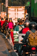 Several motorcyclists are riding together closely on a busy road. They are wearing helmets and carrying various personal items. A large truck and other vehicles can be seen in the background, contributing to a congested traffic situation. The lighting suggests it might be early morning or late afternoon.