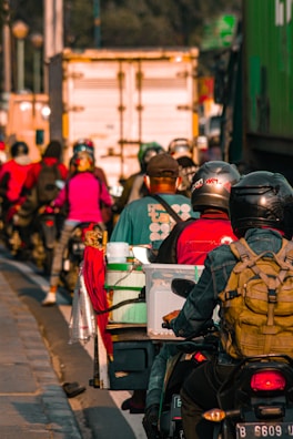 Several motorcyclists are riding together closely on a busy road. They are wearing helmets and carrying various personal items. A large truck and other vehicles can be seen in the background, contributing to a congested traffic situation. The lighting suggests it might be early morning or late afternoon.