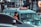 A young man in a red shirt is standing next to a light blue minivan, which has a sign on its roof reading 'JAK 43'. In the background, a driver can be seen through the van's windshield. Several vehicles, including a dark-colored car in the foreground, are also visible, along with a blurred urban setting of buildings and signage.