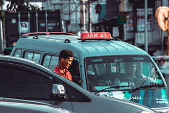 A young man in a red shirt is standing next to a light blue minivan, which has a sign on its roof reading 'JAK 43'. In the background, a driver can be seen through the van's windshield. Several vehicles, including a dark-colored car in the foreground, are also visible, along with a blurred urban setting of buildings and signage.