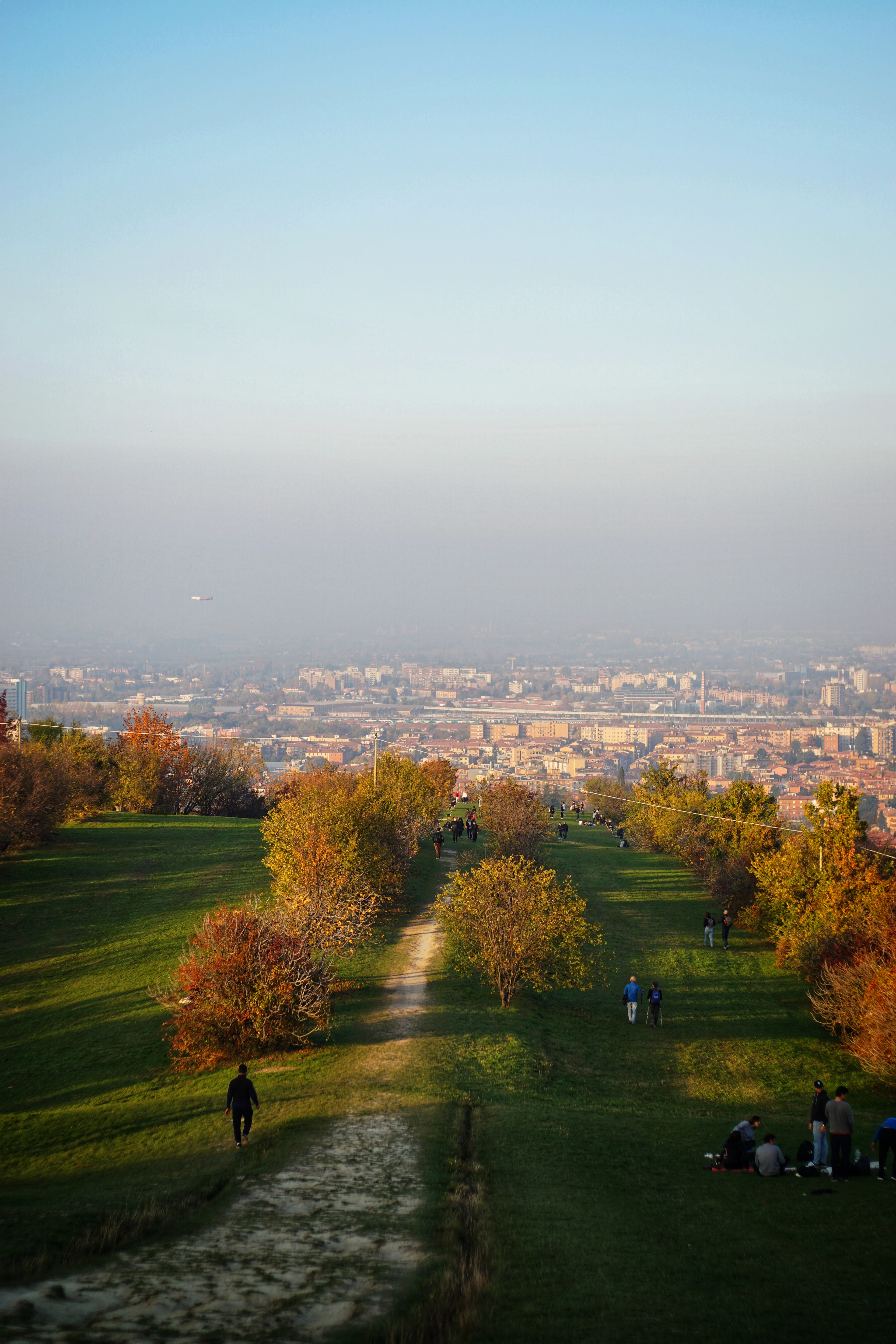 A tree-lined path over Bologna's hills leads down toward the hazy city skyline at sunset in autumn.