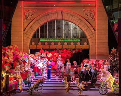 A festive scene set in front of an ornate building facade with an arched entrance, adorned with large stacks of colorful wrapped presents. Numerous figures, including children, elves, and Santa Claus, are engaged in holiday-themed activities. A decorated Christmas tree adds to the festive decoration, and a person in a bunny suit rides a bicycle near the forefront.