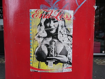 A weathered poster is affixed to a large, cylindrical red surface, featuring an illustration of a person with long hair holding a drink can. The background includes stars and a bold, artistic style with visible creases on the poster.
