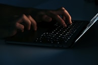Hands typing on a laptop keyboard with a holographic shield icon floating above, symbolizing cyber protection.