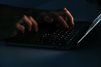 Hands typing on a laptop keyboard, illuminated by the screen's light in a dark environment.
