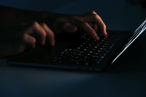 Hands typing on a laptop keyboard, illuminated by the screen's light in a dark environment.