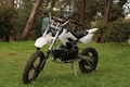 A small off-road dirt bike with a white frame and black seat is parked on a grassy area surrounded by trees. The bike features knobby tires, a robust suspension, and handlebar grips designed for rough terrain.