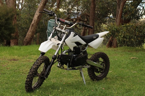 A small off-road dirt bike with a white frame and black seat is parked on a grassy area surrounded by trees. The bike features knobby tires, a robust suspension, and handlebar grips designed for rough terrain.