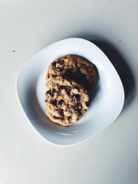 Two chocolate chip cookies are placed on a white, round plate against a light gray background. The cookies appear freshly baked with a slightly golden-brown color and visible chocolate chips.