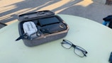 A compact drone with dark propellers is placed inside a padded carrying case on a light-colored table. Nearby, a pair of black-frame glasses rests on the table. The background includes a paved surface with shadows implying outdoor sunlight.