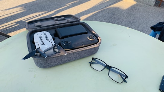 A compact drone with dark propellers is placed inside a padded carrying case on a light-colored table. Nearby, a pair of black-frame glasses rests on the table. The background includes a paved surface with shadows implying outdoor sunlight.