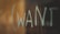 In the image, the word 'WANT' is written in light-colored chalk or marker on a transparent surface such as glass. The background is blurred, featuring warm tones, primarily shades of brown and dark colors.