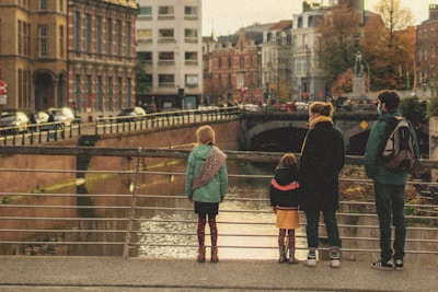 A group of four people, including two children and two adults, stand on a bridge overlooking a canal. The scene is set in an urban area with old, European-style buildings and autumn foliage visible. Each person is wearing warm clothing, suggesting cool weather.