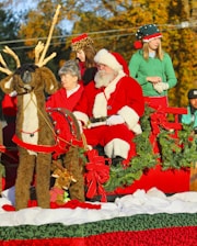 A festive holiday scene features a man in a Santa Claus costume sitting in a red sleigh adorned with greenery and large red bows. Several people accompany him, including a woman and two others dressed in Christmas-themed attire. A large plush reindeer is positioned in front of the sleigh, alongside a small stuffed reindeer wearing a red scarf. The autumn background enhances the holiday atmosphere.