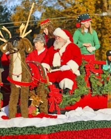 A festive holiday scene features a man in a Santa Claus costume sitting in a red sleigh adorned with greenery and large red bows. Several people accompany him, including a woman and two others dressed in Christmas-themed attire. A large plush reindeer is positioned in front of the sleigh, alongside a small stuffed reindeer wearing a red scarf. The autumn background enhances the holiday atmosphere.