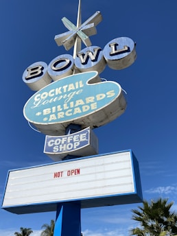 A retro-style sign advertising a bowling alley, complete with features like a cocktail lounge, billiards, arcade, and coffee shop. The sign prominently displays 'NOT OPEN' in red letters. The backdrop is a clear blue sky, and there's a hint of palm trees below.