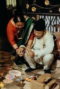 A man and a woman are participating in a traditional ceremony. The woman is wearing a green sari with a red blouse and the man is dressed in a cream-colored outfit with a cap. They are crouched near a rangoli pattern on the floor, surrounded by bricks, flowers, and ritual items like a small metal bowl.