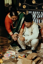 A man and a woman are participating in a traditional ceremony. The woman is wearing a green sari with a red blouse and the man is dressed in a cream-colored outfit with a cap. They are crouched near a rangoli pattern on the floor, surrounded by bricks, flowers, and ritual items like a small metal bowl.