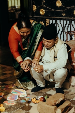 A man and a woman are participating in a traditional ceremony. The woman is wearing a green sari with a red blouse and the man is dressed in a cream-colored outfit with a cap. They are crouched near a rangoli pattern on the floor, surrounded by bricks, flowers, and ritual items like a small metal bowl.