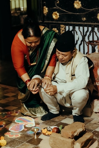 A man and a woman are participating in a traditional ceremony. The woman is wearing a green sari with a red blouse and the man is dressed in a cream-colored outfit with a cap. They are crouched near a rangoli pattern on the floor, surrounded by bricks, flowers, and ritual items like a small metal bowl.