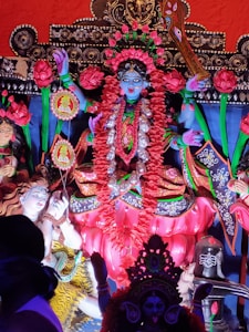 A colorful and elaborate statue depicting a Hindu goddess adorned with floral garlands, vibrant traditional attire, and intricate jewelry. The statue features detailed artistry with vivid colors, surrounded by other smaller figures and decorative elements.