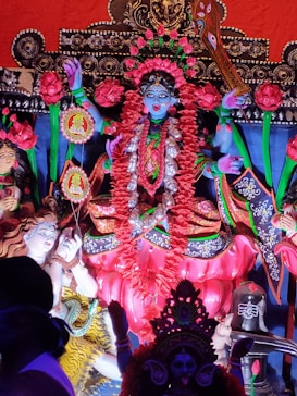 A colorful and elaborate statue depicting a Hindu goddess adorned with floral garlands, vibrant traditional attire, and intricate jewelry. The statue features detailed artistry with vivid colors, surrounded by other smaller figures and decorative elements.