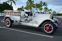 A vintage fire truck with a white body and red wheel rims is parked in an outdoor lot. The truck is labeled 'Engine No. 24' and features a mix of chrome and brass fittings, with wooden ladders and hoses on top. Palm trees in the background suggest a tropical location.