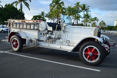 A vintage fire truck with a white body and red wheel rims is parked in an outdoor lot. The truck is labeled 'Engine No. 24' and features a mix of chrome and brass fittings, with wooden ladders and hoses on top. Palm trees in the background suggest a tropical location.