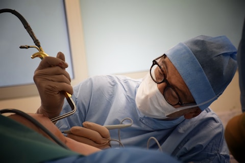 A surgeon wearing a blue surgical gown, cap, mask, and gloves is intently focused on a procedure. They are holding a surgical instrument while performing surgery. The scene is well-lit with a soft, clinical ambiance.
