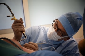 A surgeon wearing a blue surgical gown, cap, mask, and gloves is intently focused on a procedure. They are holding a surgical instrument while performing surgery. The scene is well-lit with a soft, clinical ambiance.