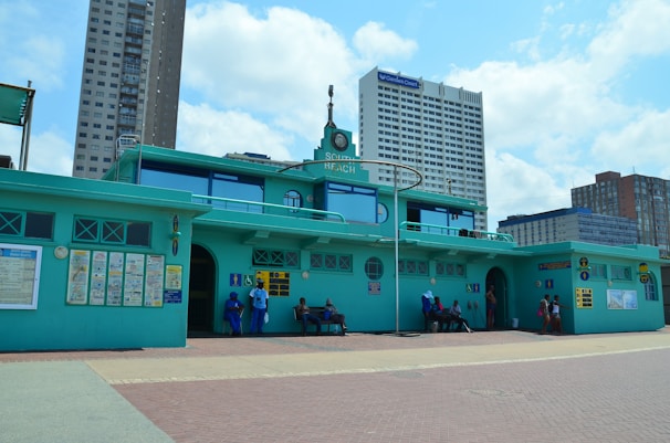 A building with a teal color scheme and a sign reading 'South Beach' is positioned in an urban setting with tall modern buildings in the background. People are seated and standing near the building, with signs and informational posters attached to the walls.
