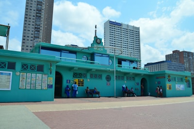 A building with a teal color scheme and a sign reading 'South Beach' is positioned in an urban setting with tall modern buildings in the background. People are seated and standing near the building, with signs and informational posters attached to the walls.