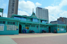 A building with a teal color scheme and a sign reading 'South Beach' is positioned in an urban setting with tall modern buildings in the background. People are seated and standing near the building, with signs and informational posters attached to the walls.