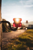 A red insulator attached to a wooden pole with the sun setting in the background, casting a warm glow. The surroundings are slightly blurred, with green grass and hints of structures in the background out of focus.