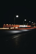 A nighttime street scene featuring a well-lit bridge or structure with modern, arched design and bright overhead streetlights. The light trails from passing vehicles create dynamic streaks across the roadway beneath. The sky is dark, complimenting the artificial lighting, while the streets are near empty.
