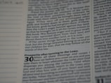 A close-up view of a page from a book with text in English. The page contains religious or biblical text, featuring bold and italicized headings along with sequential verse numbers.